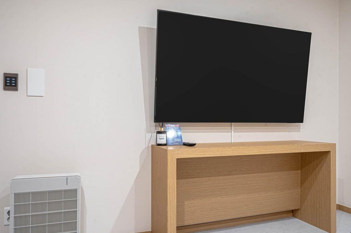 Una habitación con un televisor de pantalla plana grande montado en la pared, una mesa consola de madera, un radiador blanco y un panel de control con un control remoto.