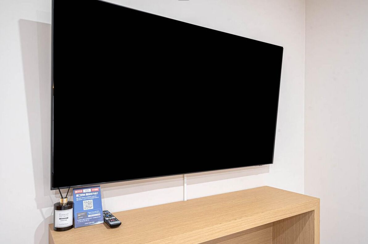 A flat-screen TV mounted on a wall with a remote control and a QR code on a wooden stand below.