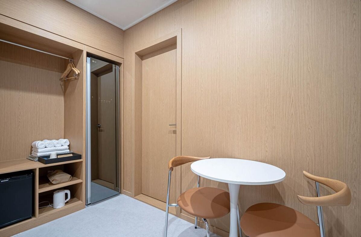 Habitación compacta con armario, una pequeña mesa y dos sillas, y una estantería con toallas y cafetera.