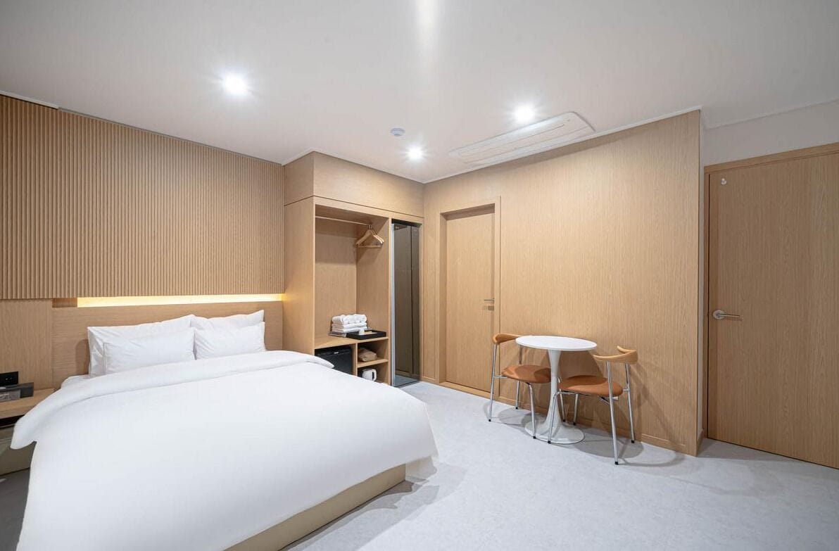 A modern hotel room with a large bed, a bedside table, a wardrobe, a seating area with a table and chairs, and a walk-in closet.