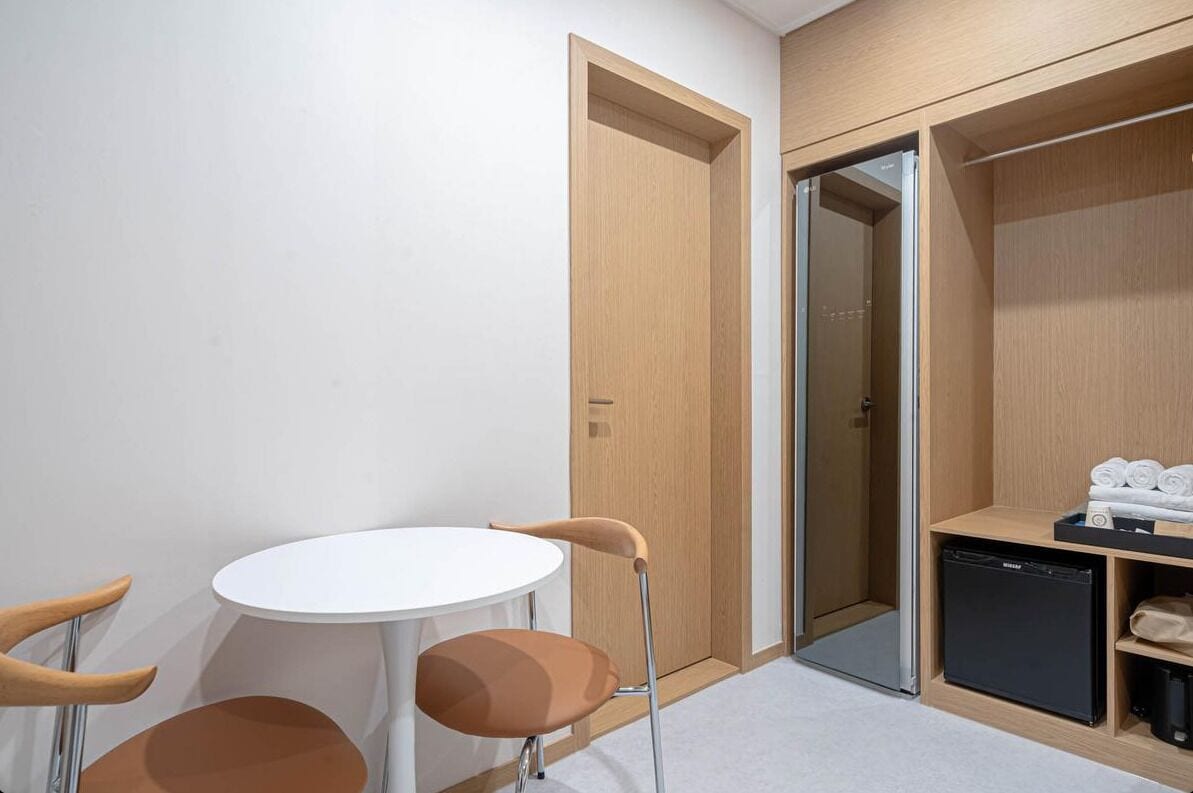 A compact room with a white table and two chairs, a wooden wardrobe with a black appliance, and a glass door leading to another room.