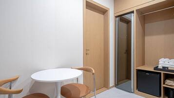 A compact room with a white table and two chairs, a wooden wardrobe with a black appliance, and a glass door leading to another room.