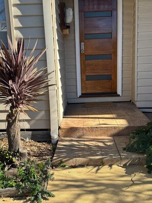 Une entrée de maison avec une porte en bois, une marche en béton et un petit jardin agrémenté d’une plante.