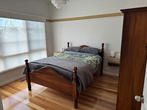 A bedroom with a wooden bed, a nightstand, a lamp, and a window with blinds. - Newport House 2 bedroom, 1 sofa bed, 1 bathroom (Newport)