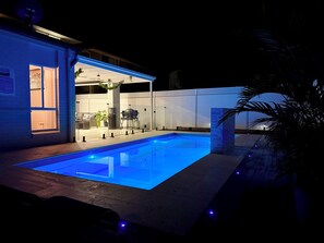 Une piscine offrant une vue dégagée sur l’intérieur du bâtiment voisin, notamment sur un espace salon et un coin barbecue.