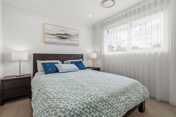 Une chambre avec un grand lit, deux tables de chevet, une commode, une fenêtre avec des stores et un tableau au mur.