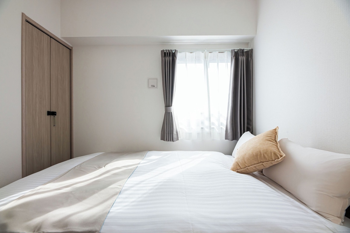 A hotel room with a bed, a wardrobe, a window with curtains, and a wall socket.
