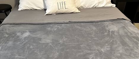 Una cama con una manta gris, dos almohadas con texto, un control remoto y una botella sobre una mesita de noche.