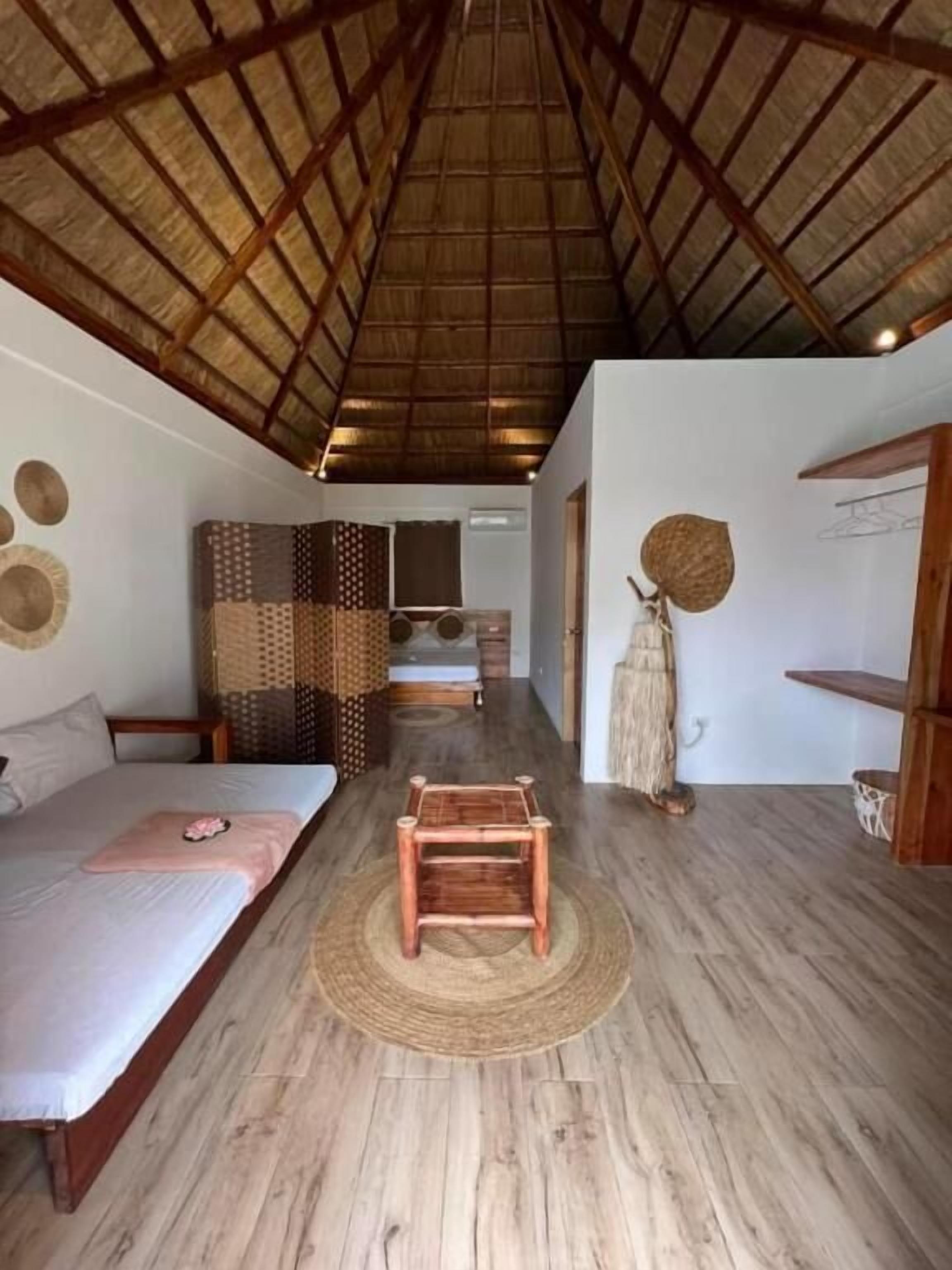 A room with a bed, a small wooden table, a woven mat, and a thatched roof ceiling.