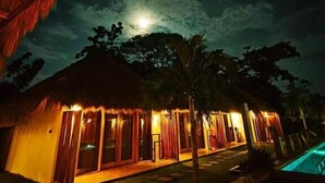 Un edificio con techo de paja y una piscina, bajo un cielo nocturno con luna visible.