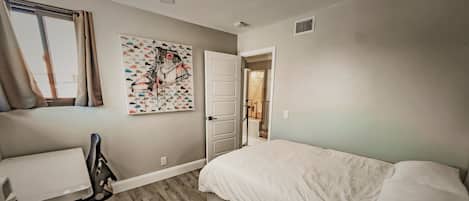 A bedroom with a bed, a desk, a chair, a window with curtains, and a wall with a shoe-themed artwork.