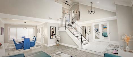 A spacious living area with a staircase, a dining area with blue chairs, and a kitchen visible in the background.