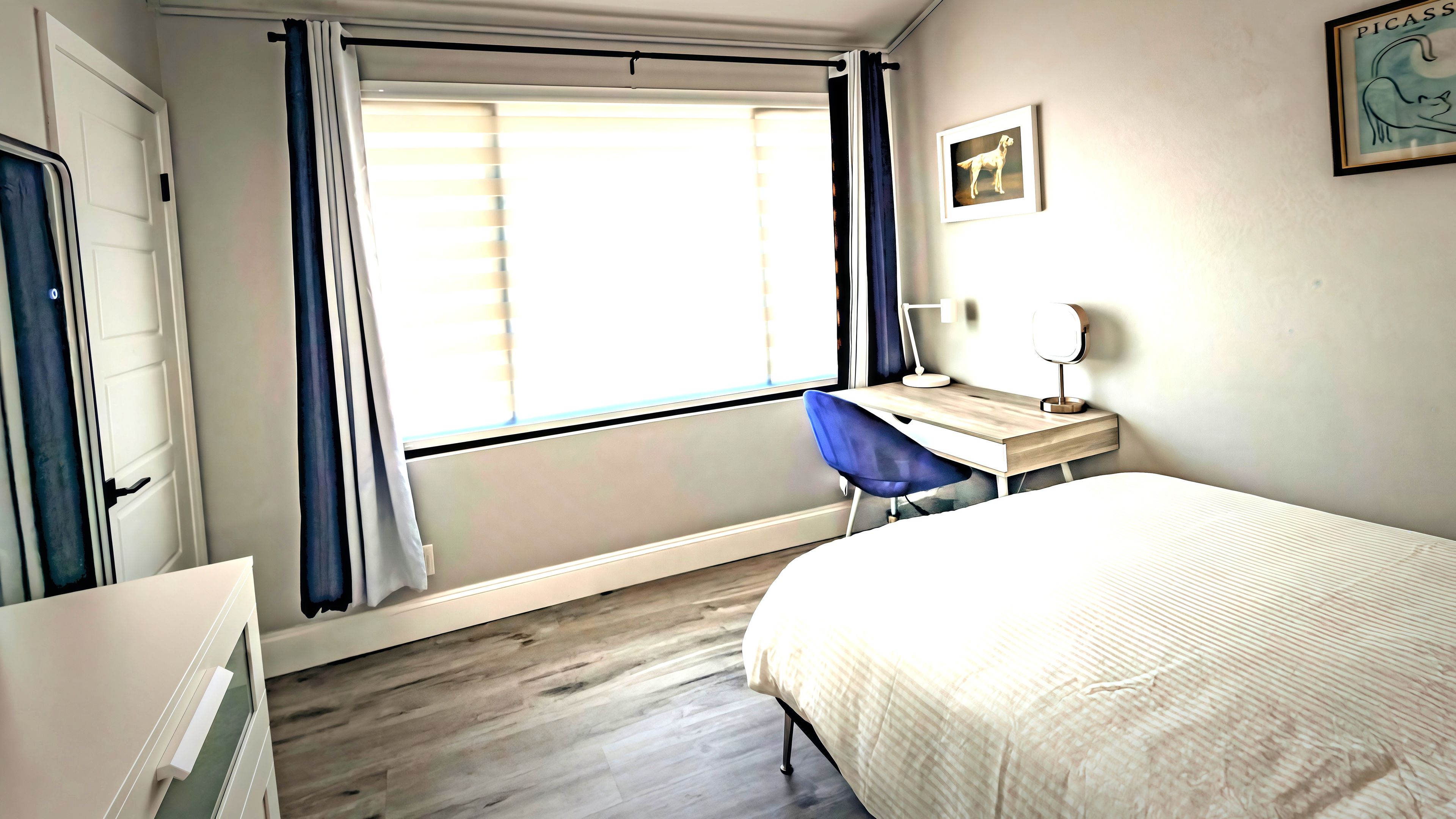 A bedroom with a bed, a desk, a chair, a window with curtains, and a framed picture on the wall.