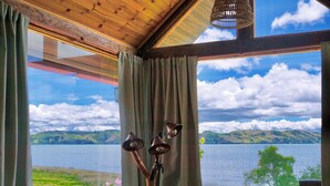 A room with a large window overlooking a lake and hills, a wooden ceiling, and a lamp with a decorative base. - The Best View in the Region, Private Beach, Cabin (Pet-Friendly) (Boyacá)