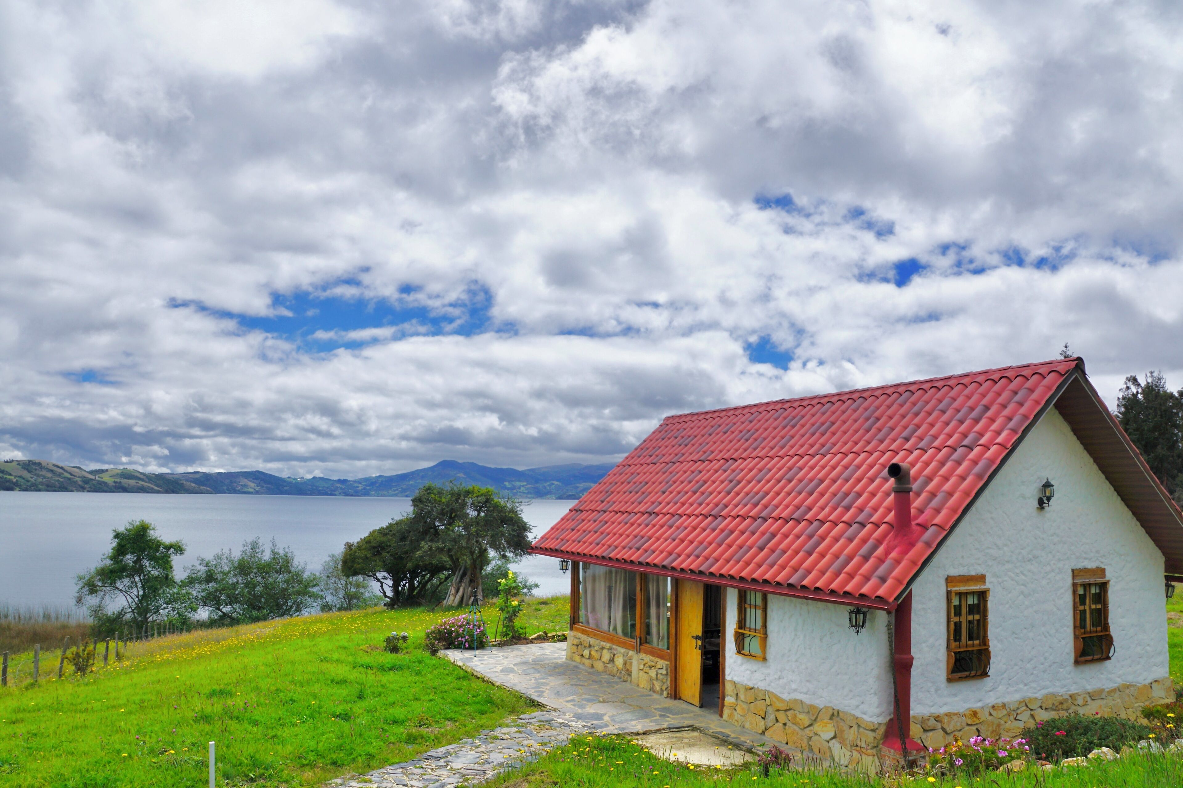 A house with a red roof, stone foundation, and a view of a lake and mountains in the background.