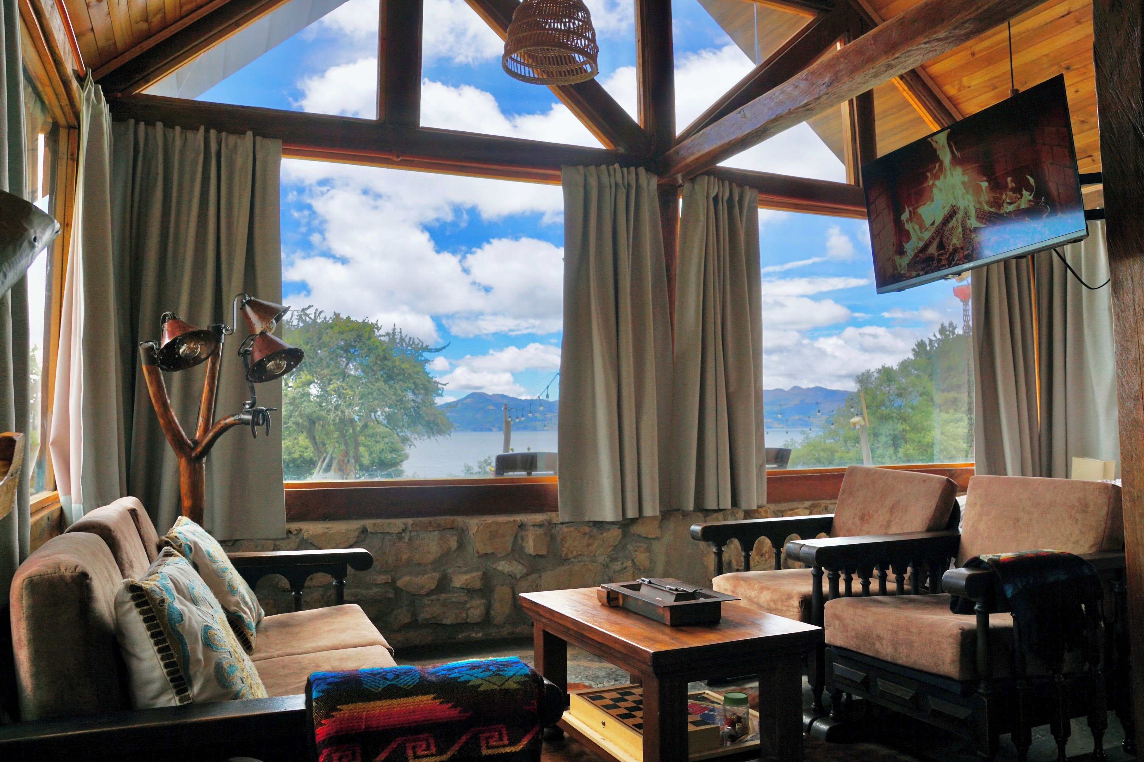 A cozy room with a stone wall, a wooden ceiling, and a large window offering a view of trees and mountains. There is a television mounted on the wall, a comfortable sofa, and a small table with a chessboard.