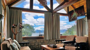 A cozy room with a stone wall, a wooden ceiling, and a large window offering a view of trees and mountains. There is a television mounted on the wall, a comfortable sofa, and a small table with a chessboard. - The Best View in the Region, Private Beach, Cabin (Pet-Friendly) (Boyacá)