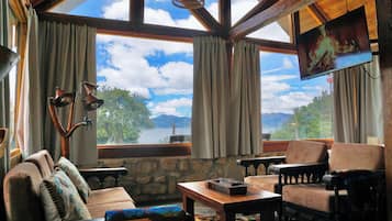 A cozy room with a stone wall, a wooden ceiling, and a large window offering a view of trees and mountains. There is a television mounted on the wall, a comfortable sofa, and a small table with a chessboard.