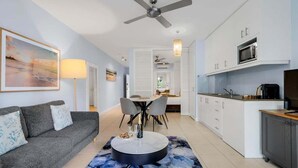 A modern living room with a sofa, a dining table, and chairs, a kitchen with white cabinets, and a ceiling fan. - 3434 Peppers Spa Penthouse (Palm Cove)
