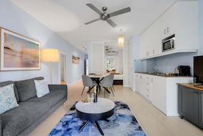 A modern living room with a sofa, a dining table, and chairs, a kitchen with white cabinets, and a ceiling fan. - 3434 Peppers Spa Penthouse (Palm Cove)