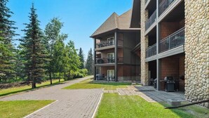 A multi-story building with balconies, a stone facade, and a paved courtyard with grass. - Chic Twin Rockies Getaway - Pool&HotTub | Sleeps 8 (Canmore)