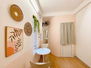 A small, well-lit room with a white round table, a white chair, and a small potted plant. There is a white door with a curtain, a wall-mounted fan, and two decorative wall hangings. - Group Stay for 4 • Big Beds • Hanoi Old Quarter Centre • Locker • Free Wash (Hà Nội)