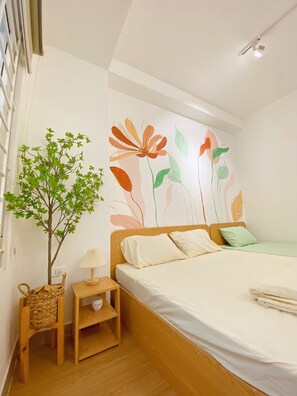 Un dormitori amb un llit de fusta, una tauleta de nit, una làmpada, una planta petita i una paret amb un mural floral.