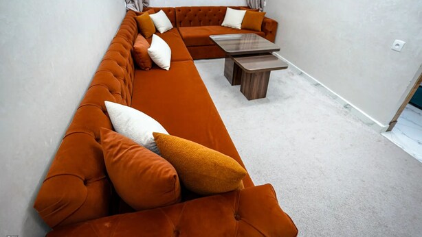 Un salon moderne avec un long canapé d’angle orange, une table basse en bois et une fenêtre avec des rideaux.