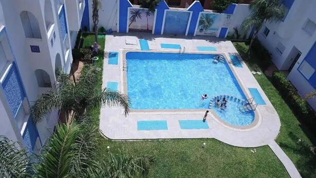 A swimming pool surrounded by a white building with blue accents, a grassy area, and palm trees. - Charming 2-bedroom Apartment With Garden View in Martil (Martil)