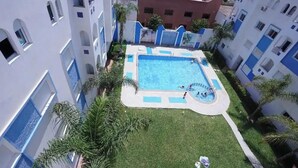 A swimming pool surrounded by a small lawn and palm trees, with buildings in the background. - Charming 2-bedroom Apartment With Garden View in Martil (Martil)