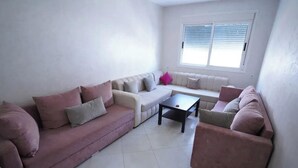 A living room with a sofa set, a coffee table, and a window with blinds. - Charming 2-bedroom Apartment With Garden View in Martil (Martil)