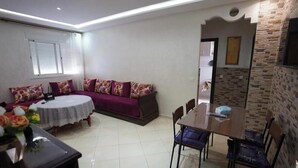 A living room with a sofa, a dining table, and chairs. There is a window with blinds, a vase with flowers, and a wall with a textured pattern. - Charming 2 -bedroom Apartment in Martil, Morocco (Martil)