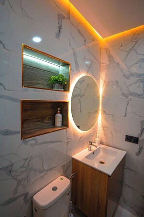A bathroom with a marble wall, a wooden shelf with a bottle, a mirror, and a toilet. - Charming 2-bedroom Apartment in the Heart of Martil, Morocco (Martil)