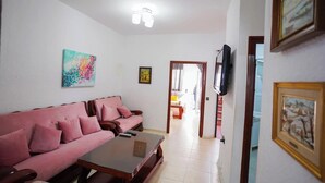 A living room with a pink sofa, a coffee table, a painting, and a television on the wall. - Charming 1-bedroom Apartment With City View in Martil (Martil)
