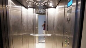 Interior de elevador com paredes metálicas, um visor digital e painel de comandos com botões.