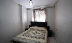 A bedroom with a bed, a nightstand, a window with curtains, and a hanging light fixture. - Charming Accessible Apartment in Martil, Morocco (Martil)