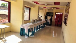 A long corridor with a row of tables covered in tablecloths, a ceiling fan, and a window with a view of a building outside. - Hotel Rama Palace (Nashik)