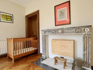 Een kamer met een houten kinderbedje, een open haard met marmeren details, en een inlijstbaar kunstwerk aan de muur.