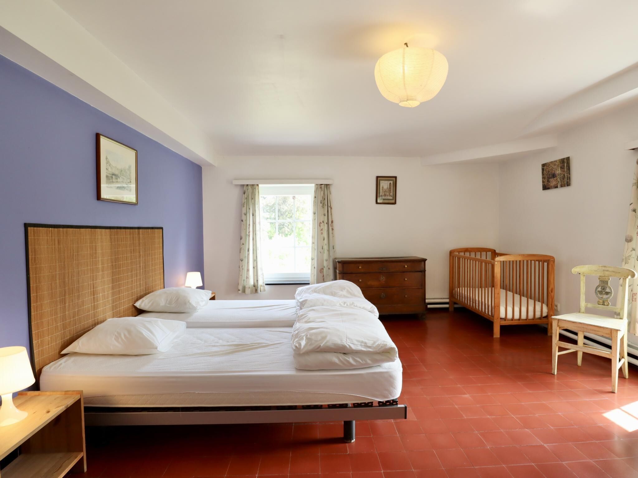 A hotel room with two beds, a wooden crib, a wooden chair, a nightstand with a lamp, and a window with curtains.