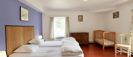 A hotel room with two beds, a wooden crib, a wooden chair, a nightstand with a lamp, and a window with curtains.