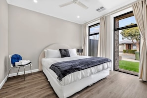 Ein modernes Schlafzimmer mit einem großen Bett, einem Sessel, einem Fenster mit Vorhängen und Blick ins Freie.