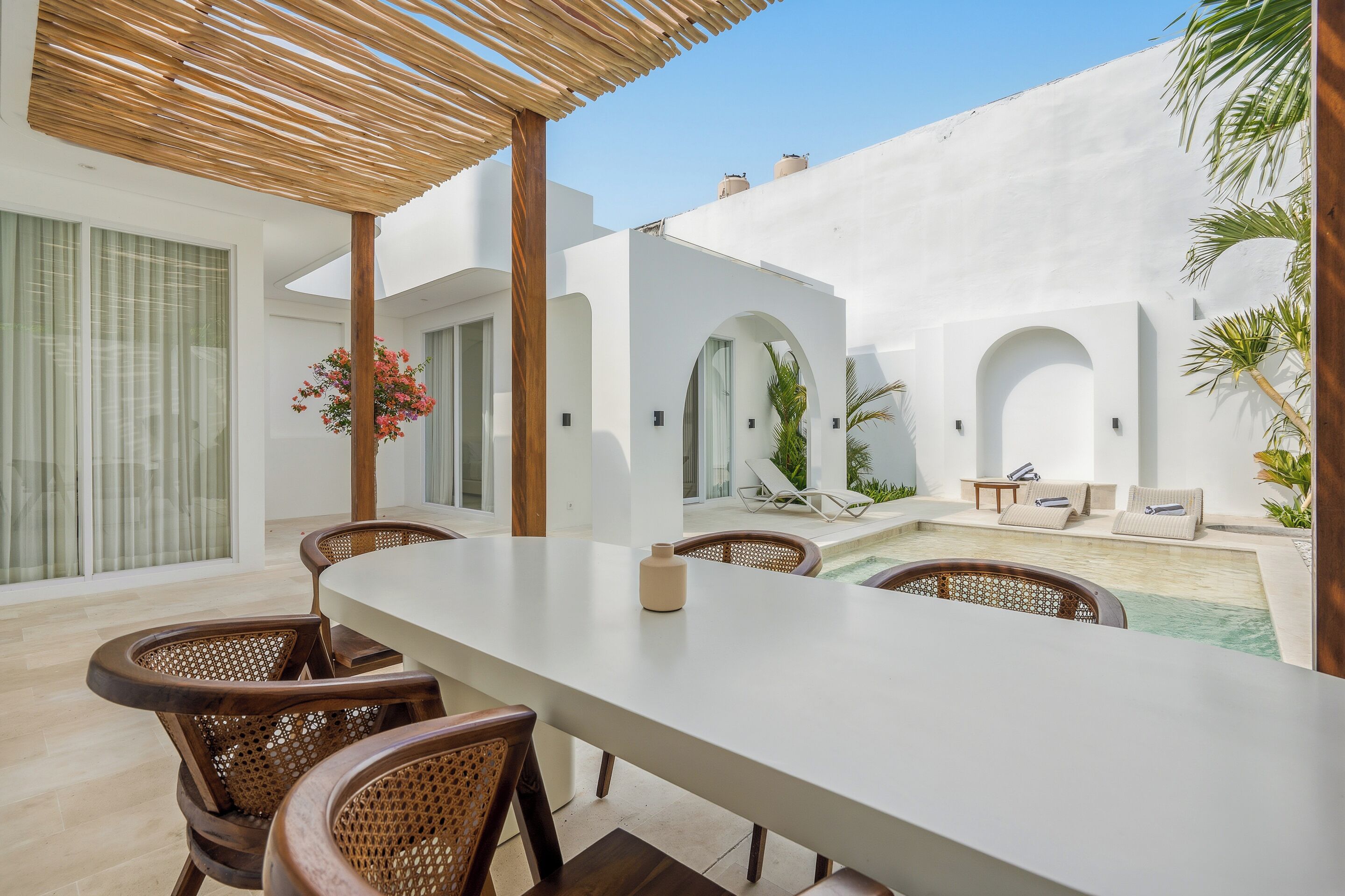 Un espacio exterior moderno para comer con una mesa blanca y sillas de mimbre, una piscina y un área de estar cubierta con una pérgola.