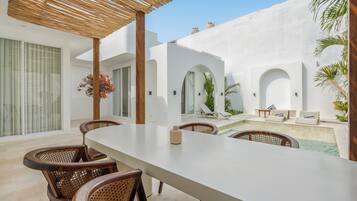 Un espacio exterior moderno para comer con una mesa blanca y sillas de mimbre, una piscina y un área de estar cubierta con una pérgola.