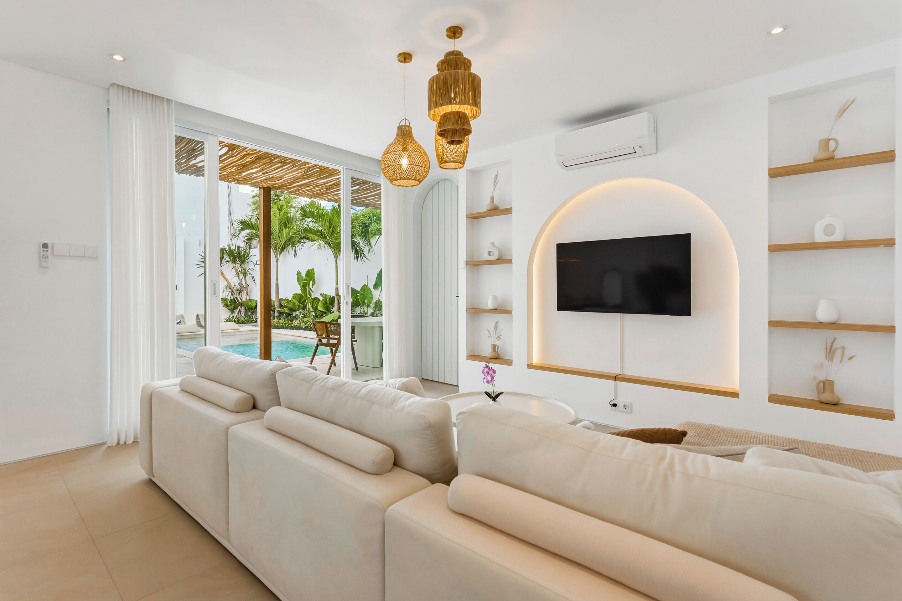 Un salon moderne avec un grand canapé d’angle, un téléviseur à écran plat fixé sur une étagère en bois, et une piscine visible à travers la porte vitrée.