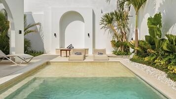 Un área de piscina con dos sillas de descanso y una mesita, rodeada de plantas tropicales y un muro blanco.