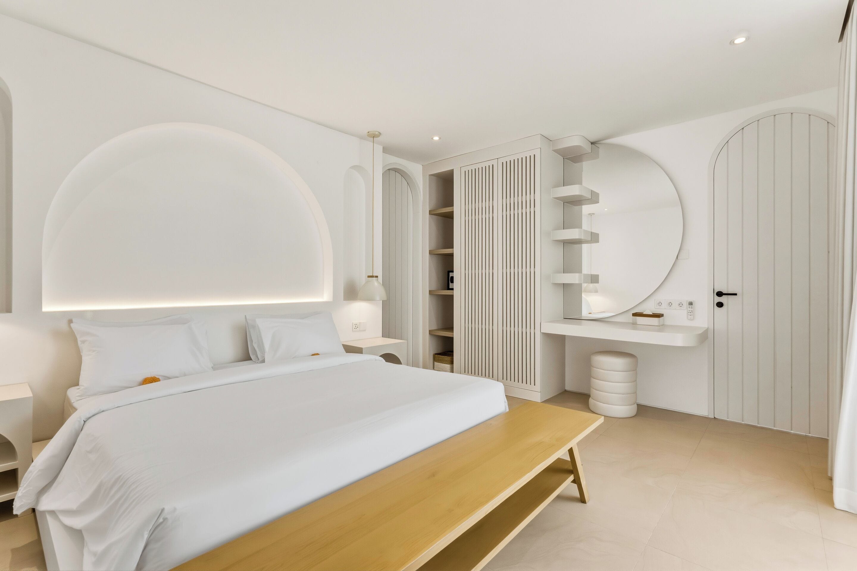 Un dormitorio moderno con una cama grande, mesitas de noche, un banco empotrado, un armario, un espejo y un espejo redondo en la pared.