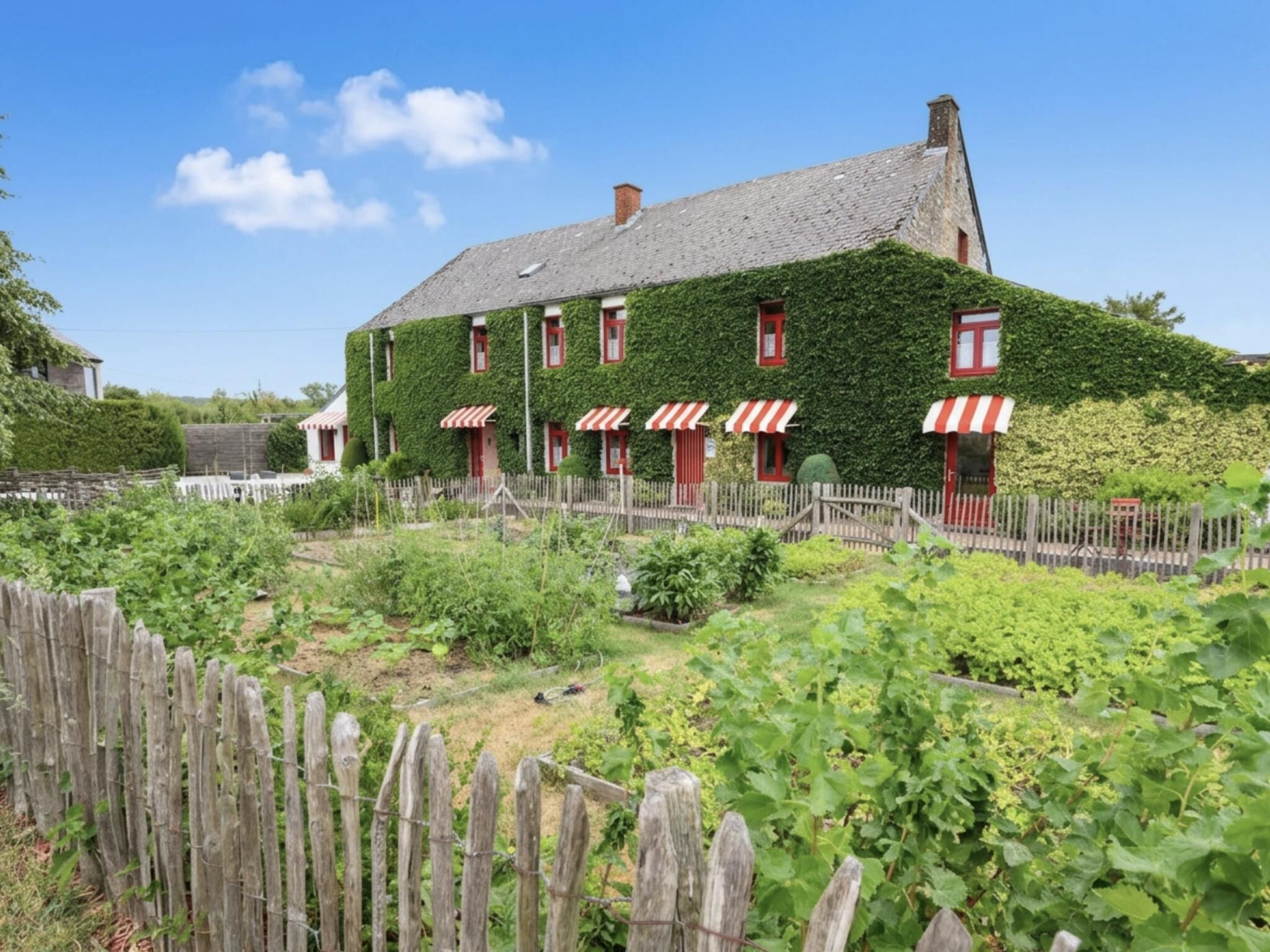 Un bâtiment doté d’un toit en pente, d’auvents à rayures rouges et blanches et d’un jardin où poussent différentes espèces de plantes.