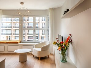 A modern living room with a sofa, a round table, a vase with flowers, and a view of a building through large windows. - Stylish apartment with dune view near Idesbald (Sint-Idesbald)