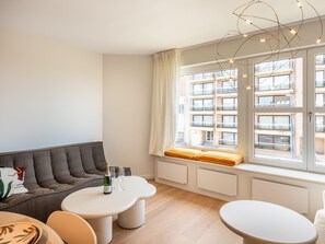 Ein modernes Wohnzimmer mit einer Couch, zwei weißen Tischen und einem großen Fenster mit Blick auf Gebäude.