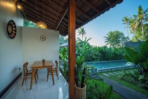 A covered patio with a dining table and chairs, a hanging light fixture, and a view of a swimming pool surrounded by lush greenery. - Wahana Cottage Ubud (Gianyar)